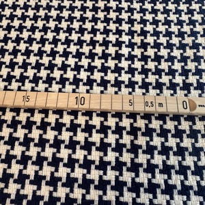 May include: Close-up of a houndstooth fabric in navy blue and cream. The fabric has a woven texture and a classic pattern. A wooden ruler with metric measurements is placed across the fabric.