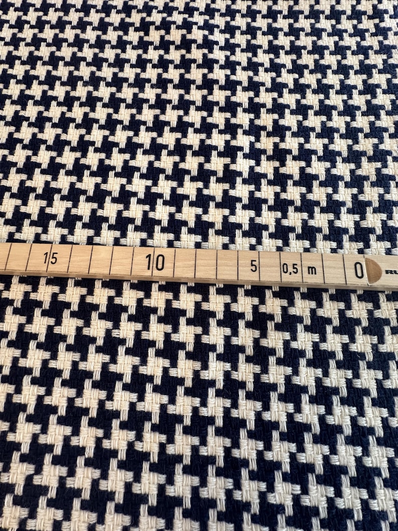 May include: Close-up of a houndstooth fabric in navy blue and cream. The fabric has a woven texture and a classic pattern. A wooden ruler with metric measurements is placed across the fabric.