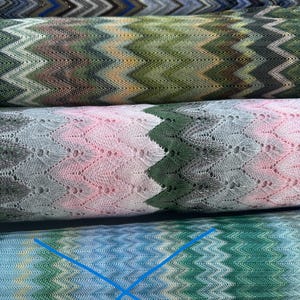 Könnte beinhalten: Vier Rollen Strickstoff mit einem sich wiederholenden Zickzack-Chevron-Muster. Die oberste Rolle ist blau und braun, die zweite grün und braun, die dritte rosa und grau mit Lochmuster und die untere Rolle ist blau und grün mit einem blauen X.