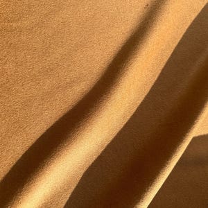 May include: Close-up of a tan fabric with a soft, textured surface. The fabric is draped, with shadows creating diagonal lines across the surface. The color is a warm, earthy tone, suggesting a natural material.
