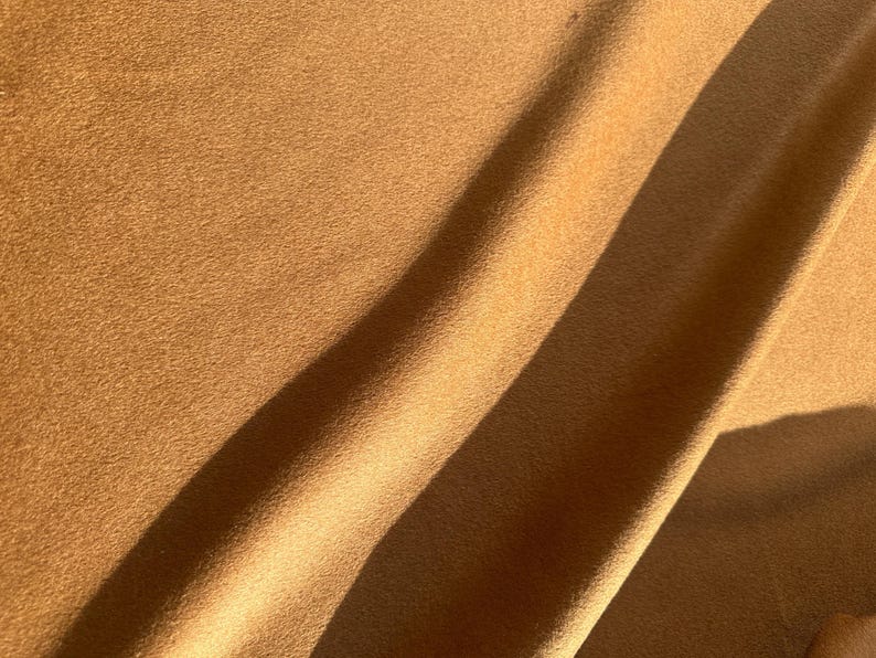 May include: Close-up of a tan fabric with a soft, textured surface. The fabric is draped, with shadows creating diagonal lines across the surface. The color is a warm, earthy tone, suggesting a natural material.