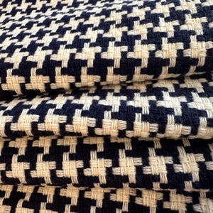 May include: Close-up of folded fabric with a woven houndstooth pattern. The fabric features a repeating design of navy blue and off-white squares. The texture appears soft and the fabric is folded over itself.