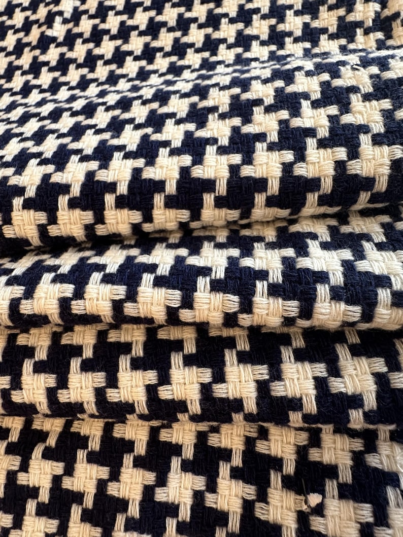 May include: Close-up of folded fabric with a woven houndstooth pattern. The fabric features a repeating design of navy blue and off-white squares. The texture appears soft and the fabric is folded over itself.