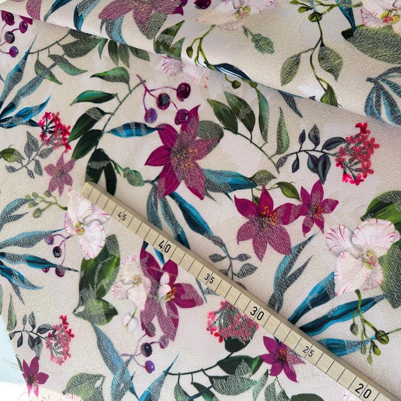 May include: A close-up of a cream-colored fabric with a floral print. The fabric features pink, purple, and white flowers with green leaves. A measuring tape is visible in the bottom right corner of the image.