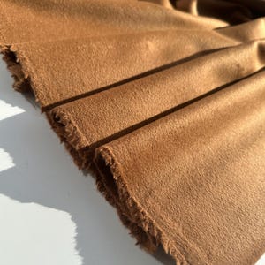 May include: Close-up of a folded piece of brown fabric with a soft, textured surface. The edges of the fabric are frayed, revealing the raw texture. The fabric is a warm, earthy tone, with visible folds and shadows.