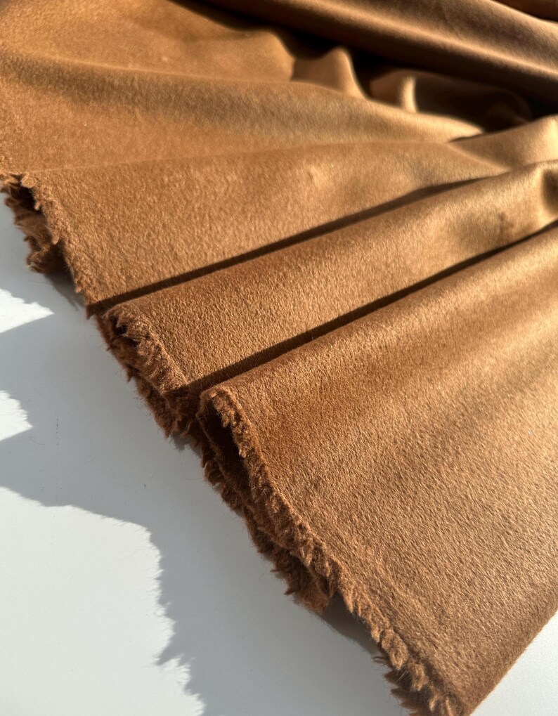 May include: Close-up of a folded piece of brown fabric with a soft, textured surface. The edges of the fabric are frayed, revealing the raw texture. The fabric is a warm, earthy tone, with visible folds and shadows.