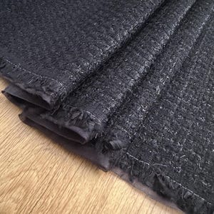 May include: Close-up of a dark navy fabric with a textured weave. The fabric has a subtle sheen and is folded, revealing a layered construction. The edges are finished with a delicate fringe.