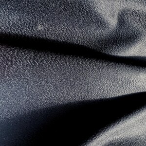 May include: A dark charcoal gray fabric with a fine, dense, and slightly pebbled texture. The material is draped to create deep, soft folds and shadows, highlighting the subtle grain and matte finish of the textile surface across the frame.