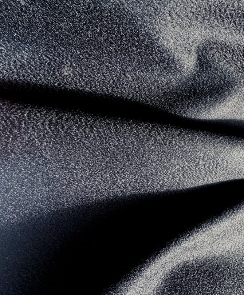 May include: A dark charcoal gray fabric with a fine, dense, and slightly pebbled texture. The material is draped to create deep, soft folds and shadows, highlighting the subtle grain and matte finish of the textile surface across the frame.