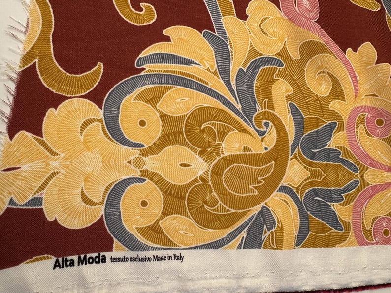 May include: A close-up of a fabric with a paisley pattern in shades of brown, yellow, blue, and pink. The fabric has a white border with the text "Alta Moda tessuto esclusivo Made in Italy".