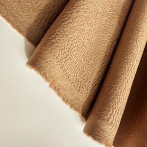May include: A piece of tan fabric with a distinct, raised, and wavy textured surface. The material is draped in soft, vertical folds, showcasing the consistent light brown color and the intricate, rippled pattern across the entire textile.