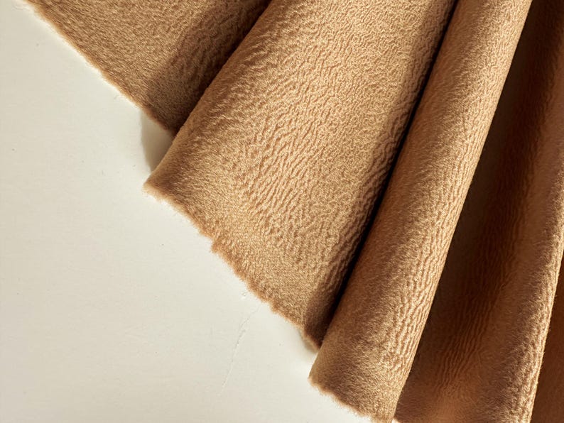 May include: A piece of tan fabric with a distinct, raised, and wavy textured surface. The material is draped in soft, vertical folds, showcasing the consistent light brown color and the intricate, rippled pattern across the entire textile.