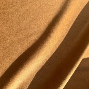 May include: A solid tan-colored fabric with a soft, matte, and slightly fuzzy texture. The material is draped in gentle folds, creating diagonal shadows across the surface that highlight the consistent, warm brown tone and the fine, uniform nap.
