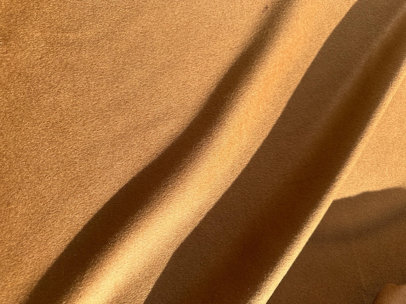 May include: A solid tan-colored fabric with a soft, matte, and slightly fuzzy texture. The material is draped in gentle folds, creating diagonal shadows across the surface that highlight the consistent, warm brown tone and the fine, uniform nap.