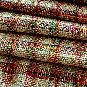 Puede incluir: Primer plano de una tela tejida con un fondo beige e hilos coloridos tejidos en un patr&oacute;n de cuadros. La tela est&aacute; doblada en capas, mostrando la textura y el detalle del tejido.