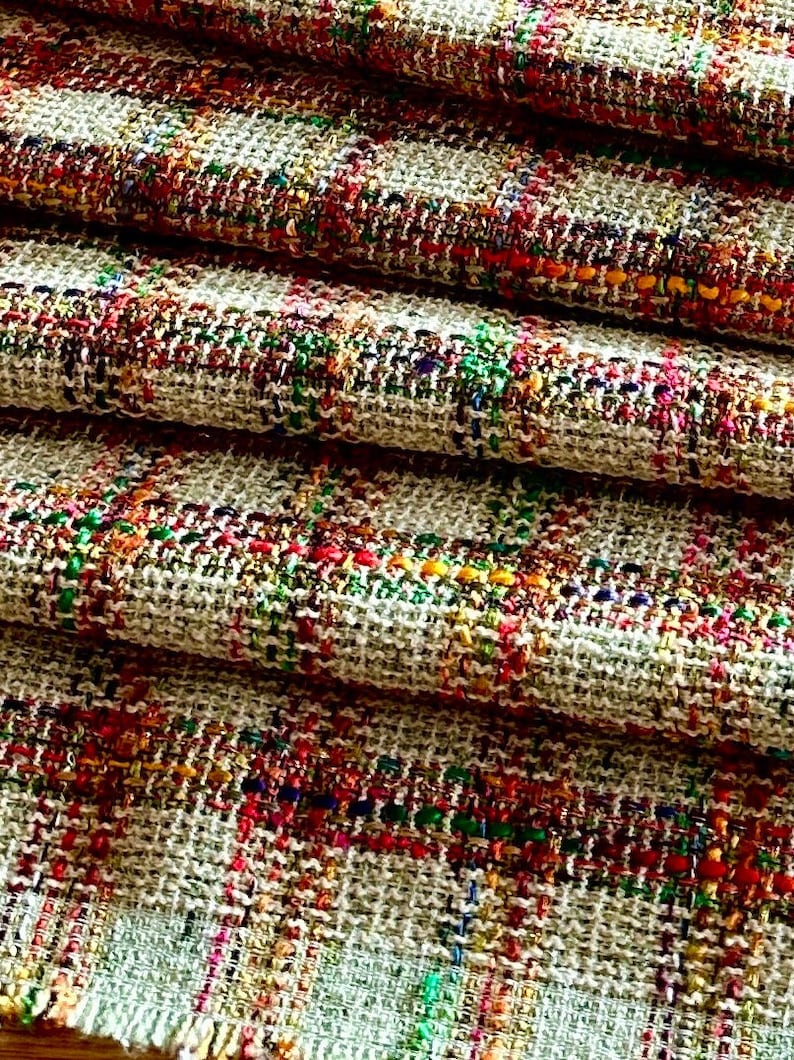 Puede incluir: Primer plano de una tela tejida con un fondo beige e hilos coloridos tejidos en un patr&oacute;n de cuadros. La tela est&aacute; doblada en capas, mostrando la textura y el detalle del tejido.