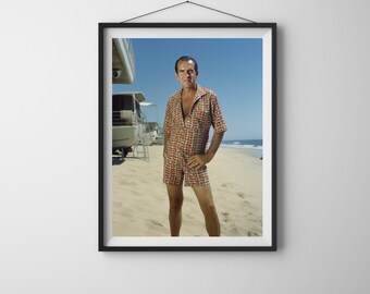May include: A man wearing a red and white patterned short-sleeved shirt and matching shorts stands on a sandy beach. He is looking at the camera with a serious expression.