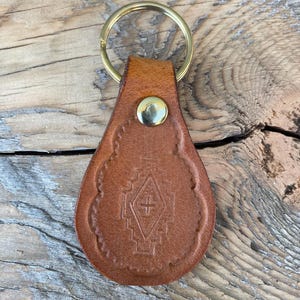 May include: A brown leather keychain with a gold key ring and a decorative design featuring a cross inside a geometric pattern.
