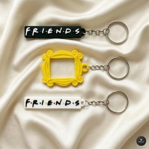 Könnte beinhalten: Drei Schlüsselanhänger mit dem Wort "FRIENDS" und einem gelben Rahmen. Ein Schlüsselanhänger ist schwarz mit weißen Buchstaben, ein anderer ist weiß mit bunten Buchstaben, und der dritte ist ein gelber Rahmen. Jeder Schlüsselanhänger hat eine silberne Kette und einen Ring.