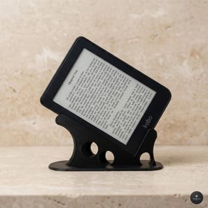 Può includere: Un e-reader Kobo nero è esposto su un supporto nero. Lo schermo mostra il testo del capitolo "Chapter Ten". Il supporto ha un design unico con tre fori circolari. Il logo Kobo è visibile.