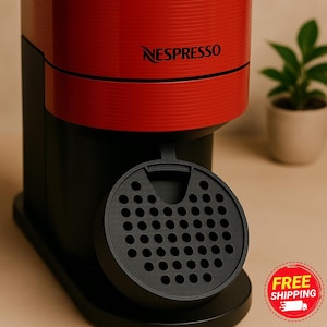 Op de afbeelding: Een rode Nespresso koffiemachine met een zwarte druppelbak en een zwarte, afneembare druppelbakdeksel met een cirkelvormig patroon van gaten. De machine heeft het woord "NESPRESSO" op de voorkant.
