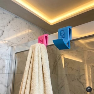 Peut inclure: Un crochet rose vif et un crochet bleu vif à suspendre sur une porte, chacun arborant un logo carré en relief sur le devant. Le crochet rose soutient une serviette beige pliée, tandis que le crochet bleu est vide. Fixés sur une porte de douche.