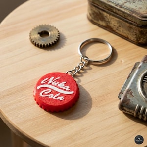 Peut inclure: Un porte-clés rouge en forme de bouchon de bouteille Nuka Cola avec du texte blanc. Le porte-clés est sur une chaîne et un anneau argentés. D'autres objets sur une surface en bois incluent un petit engrenage et une boîte en métal.