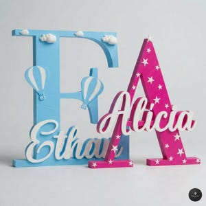 Può includere: Due lettere di legno, "E" in azzurro con dettagli di mongolfiere e nuvole, e "A" in rosa con un motivo a stelle bianche. I nomi "Ethan" e "Alicia" sono scritti in corsivo bianco.