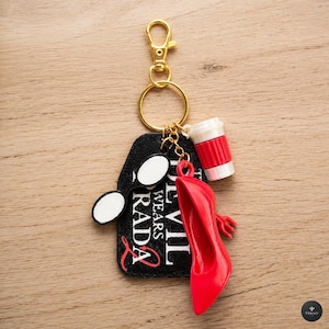 Può includere: Un portachiavi color oro con un'etichetta nera con la scritta "Devil Wears Prada" in bianco. Sono attaccati occhiali da sole bianchi, un tacco alto rosso e un ciondolo a forma di tazza di caffè rosso e bianco. Su una superficie di legno.