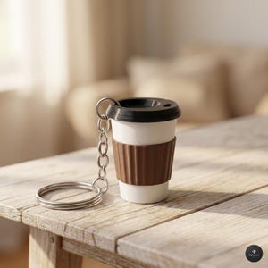 Könnte beinhalten: Ein Miniatur-Kaffeebecher-Schlüsselanhänger mit weißem Korpus, brauner Hülle und schwarzem Deckel. Der Schlüsselanhänger hat eine silberne Kette und einen Ring. Der Becher steht auf einer Holzoberfläche.