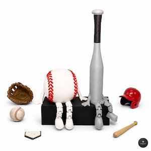 Op de afbeelding: Een witte honkbal met rode stiksels en een grijze honkbalknuppel, beide met beweegbare ledematen, zitten op een zwart rechthoekig blok. Accessoires zijn onder meer een bruine leren handschoen, een kleine honkbal, een honkplaat, een rode helm en een miniatuurknuppel.
