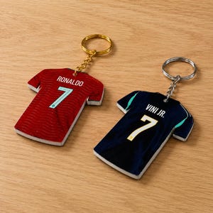 Könnte beinhalten: Zwei trikotförmige Schlüsselanhänger mit Metallringen. Der linke Anhänger ist rot mit Wellenmuster, dem Namen RONALDO und der Nummer 7 in Hellblau. Der rechte Anhänger ist dunkelblau mit dem Namen VINI JR. und der Nummer 7 in Gelb.