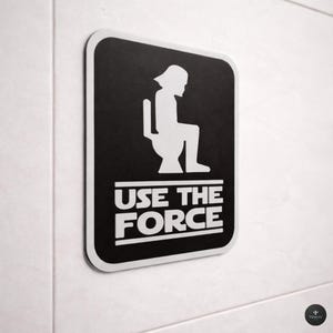 Könnte beinhalten: Ein rechteckiges schwarzes Schild mit abgerundeten Ecken und einem weißen Rand. Das Schild zeigt eine weiße Grafik einer Figur, die auf einer Toilette sitzt, über dem weißen Text USE THE FORCE, der zwischen zwei horizontalen weißen Linien steht.
