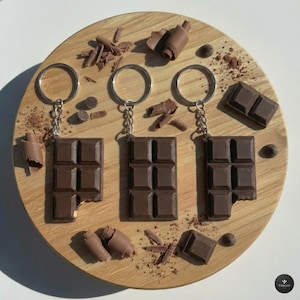 Op de afbeelding: Drie donkerbruine sleutelhangers in de vorm van chocoladerepen met zilverkleurige metalen ringen en kettingen. Elke sleutelhanger heeft een rechthoekig ontwerp met zes segmenten. Ze liggen op een licht houten oppervlak met chocoladeschaafsel.