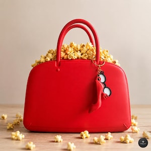 Może przedstawiać: Jasnoczerwona torebka wypełniona popcornem. Torebka ma dwa zaokrąglone uchwyty i mały brelok przedstawiający but na obcasie i okulary. Ziarna popcornu są rozrzucone wokół podstawy torebki.