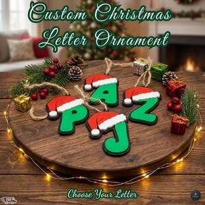 Puede incluir: Adornos de letras navideñas verdes con gorros de Papá Noel rojos y ribetes blancos. Las letras se muestran sobre una superficie de madera con decoraciones festivas, incluyendo pequeños regalos envueltos y piñas. El texto "Choose Your Letter" también es visible.
