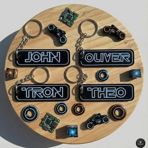 May include: A wooden display with keychains bearing the names "JOHN", "OLIVER", "TRON", and "THEO" in white on black. The display also features glowing geometric shapes and motorcycle charms, all arranged on a circular wooden base.