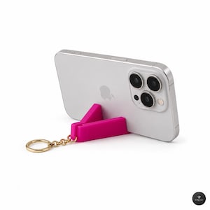 May include: A bright pink, V-shaped plastic phone stand attached to a gold-toned metal keyring. The stand is propped against the back of a silver smartphone, which features a triple-lens camera system and a centred apple logo on its rear surface.