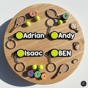Può includere: Una targa rotonda in legno con decorazioni a tema tennis. Presenta portachiavi con nomi come "Adrian", "Andy", "Isaac" e "BEN", mini racchette da tennis, palline da tennis e rotoli di nastro colorato. La targa ha una venatura del legno chiara.