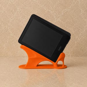 Op de afbeelding: Een zwarte Kobo e-reader staat onder een hoek in een feloranje, gebogen kunststof standaard. De standaard heeft een platte voet, een ondersteunende rugleuning en een klein cirkelvormig uitgesneden detail aan de rechterkant voor het apparaat.