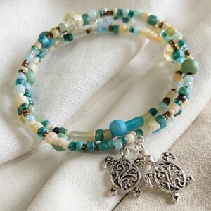 May include: A beaded wrap bracelet in shades of blue, green, and cream. The bracelet features small, colorful beads and two silver turtle charms. The bracelet is approximately 7 inches in circumference.
