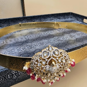May include: A gold belt with a large, ornate peacock design. The peacock is adorned with clear and white stones, and the belt has a dangling chain of red and white beads.