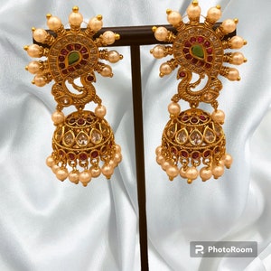 May include: A pair of gold tone earrings with a peacock design. The earrings feature a large, round, clear stone surrounded by smaller stones. The peacock design is accented with red and green stones. The earrings are suspended from a chain of pearls.