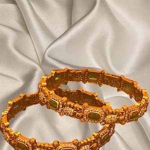 May include: Two gold bangles with intricate designs and green and pink gemstones. The bangles are on a white satin background.