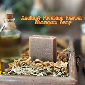 May include: A square, brown herbal shampoo soap bar sits on a bed of dried herbs and flowers. A glass bottle with a cork stopper is in the background. The text "Ancient Formula Herbal Shampoo Soap" is displayed in orange.