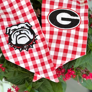 May include: Red and white gingham dog bandana with a black and white bulldog patch and a patch with a black "G" inside a black and white oval. The bandana is surrounded by green leaves and red flowers.