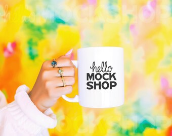 May include: White ceramic mug with black text that reads "hello mock shop". The mug is being held by a hand wearing rings.