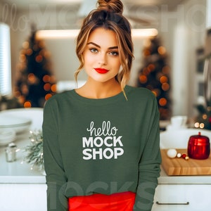 May include: A woman wearing a dark green long-sleeved shirt with the text "hello mock shop" in white. She is standing in a kitchen with Christmas trees in the background.