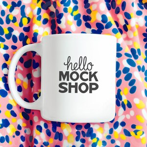May include: White ceramic mug with a black inscription that reads "hello mock shop" on a pink and blue patterned background.