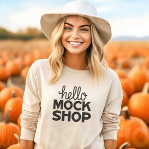 May include: A woman wearing a white hat and a beige sweatshirt with the text "hello mock shop" in black lettering. She is standing in a field of pumpkins.